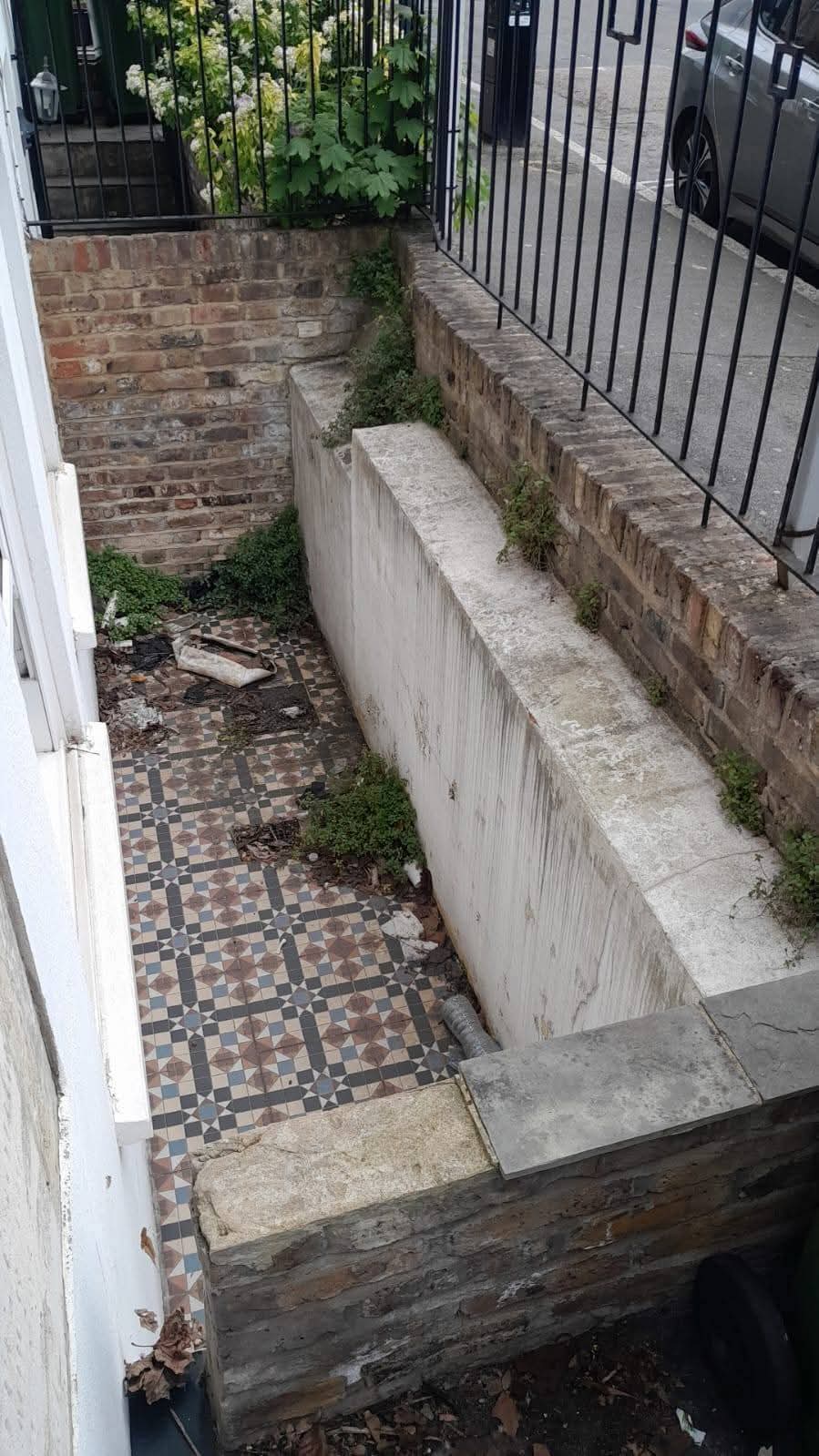 A basement lightwell with weeds and rubbish on the tiles